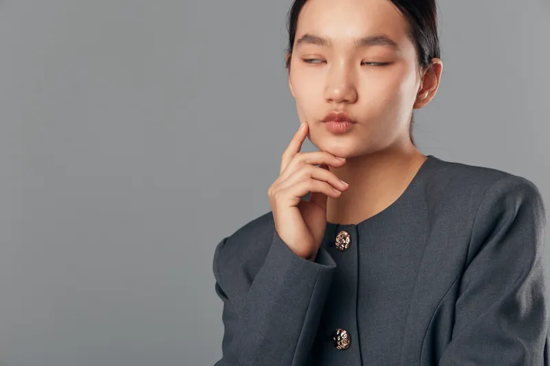Thoughtful woman with closed eyes and hand near chin in gray jacket, studio portrait on grey background. Calm, pensive expression and formal outfit for business or lifestyle concept.