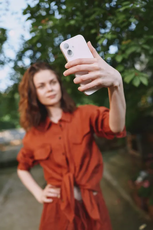 Young woman in a rust orange dress takes a selfie outdoors, capturing a casual lifestyle moment with a bright smile, green trees, and soft daylight as the focus and natural background
