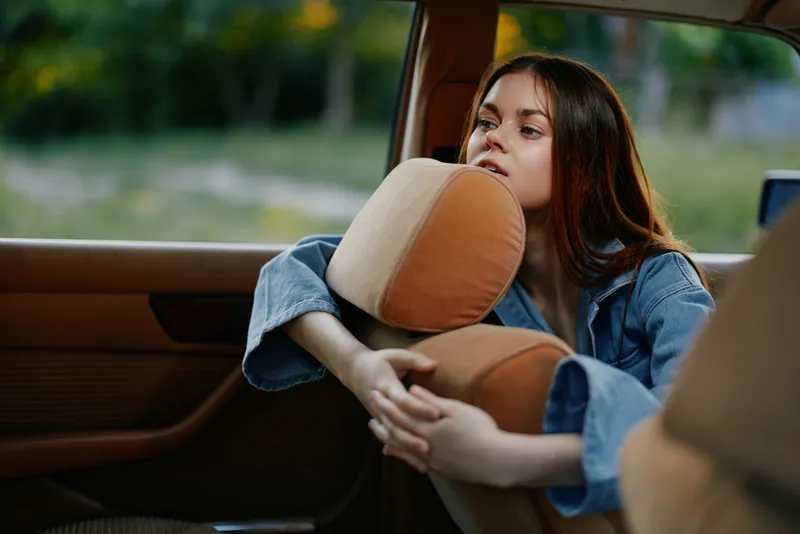 Pensive young woman in denim jacket resting on car seat, reflecting on life, with green nature background highlighting a moment of introspection and solitude.