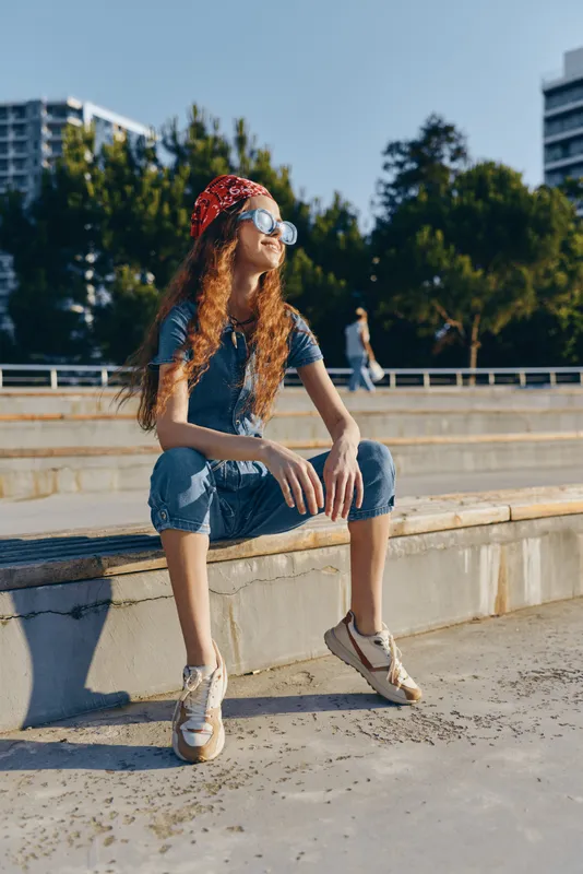 young woman with curly hair sits outdoors in casual summer clothes wearing sunglasses and a red bandana, enjoying sunny day in urban park setting
