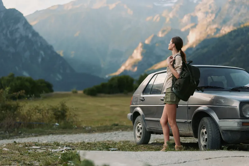 Young woman with backpack standing near car on mountain road, enjoying nature and fresh air. Concept of travel, adventure, and outdoor lifestyle in scenic environment.