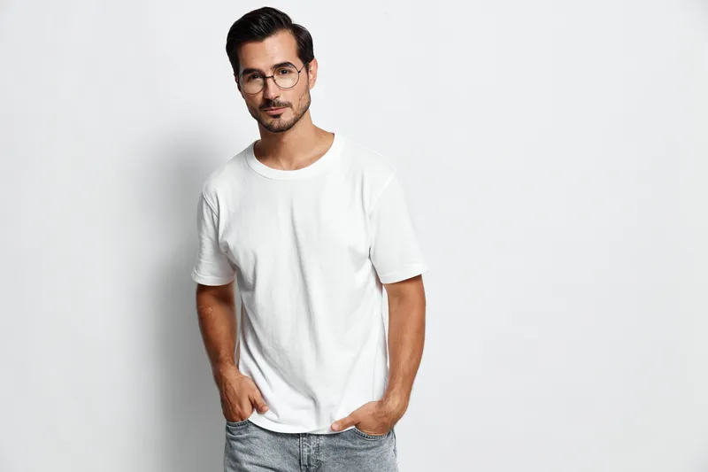 Stylish young man wearing a white t-shirt and glasses stands confidently with hands in pockets against a plain gray background. Fashion lifestyle concept.