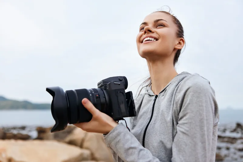 Young woman enjoying photography outdoors on a sunny day, holding a camera with a joyful expression, showcasing passion for nature and creativity