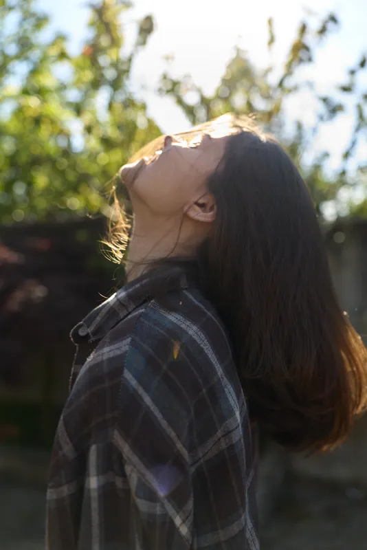 Cheerful woman outdoors, head tilted back, sunlight on her face, enjoying a relaxed lifestyle moment, plaid shirt, natural colors, soft breeze, lush greenery background, calm, candid vibe