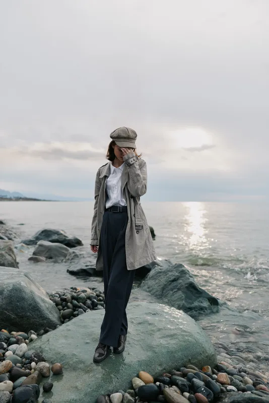 Woman in stylish coat and hat stands on rocky beach near calm sea during cloudy day. Fashion concept with casual autumn outfit by the water. Moody natural light and relaxed atmosphere.