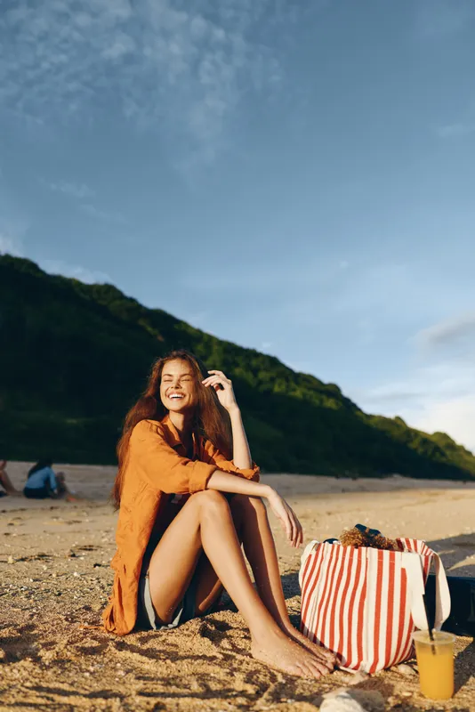 A young woman in an orange cover-up sits on sunlit beach sand, smiling with eyes closed, striped bag beside her and a refreshing drink nearby, mountains rising in the background