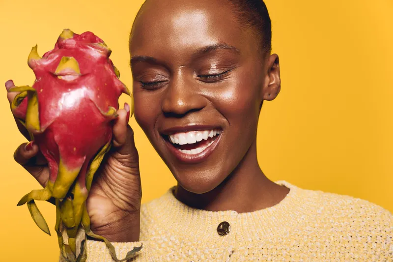 Smiling African woman with dark skin holding a red dragon fruit on yellow background. Concept of healthy lifestyle, nutrition, exotic fruit, and natural beauty with vibrant colors.