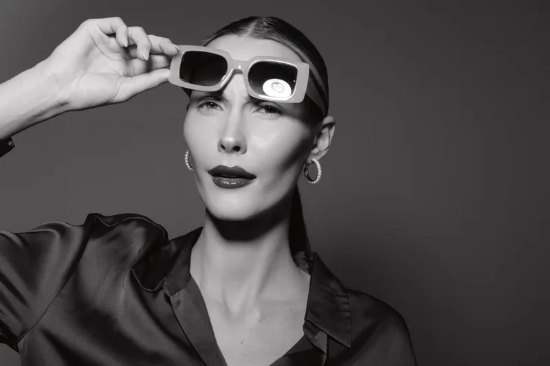 Stylish Caucasian woman with light skin adjusting sunglasses, wearing dark blouse and hoop earrings in studio. Concept for fashion, elegance, and confident attitude.