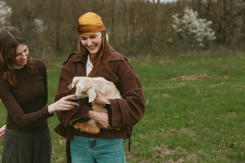 Two women in casual coats share a moment outdoors as one holds a fluffy goat kid, smiling warmly, while the other offers a gentle touch on its head in a grassy field.