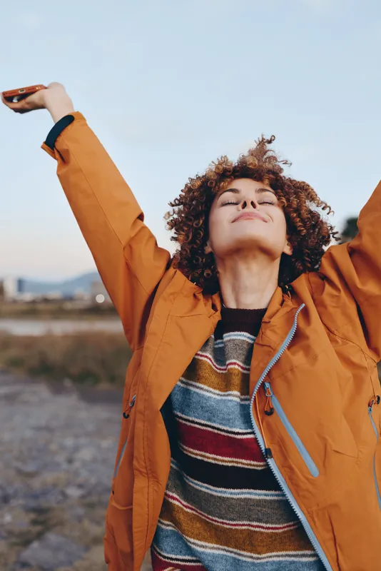woman lifestyle outdoors, curly hair, orange jacket, arms raised, looking upward at blue sky, happy smile, calm relaxed pose, street scenery and soft daylight, concept freedom and wellbeing