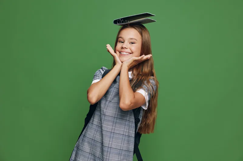 Girl smiling and holding books on head with hands near face in happy gesture for education, learning and youth on green background. Cute student, childhood and school concept.