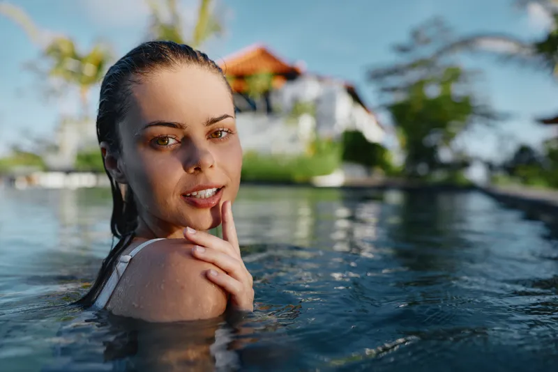 A relaxed woman enjoys a pool swim, shoulders above water, sunlit skin and a gentle smile, with a resort in the background creating a carefree lifestyle moment.