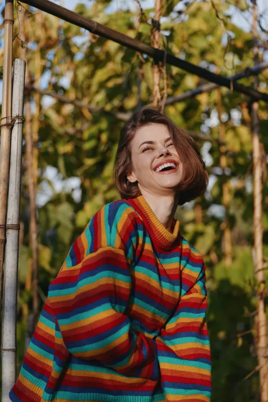 Woman smiling in a colorful rainbow sweater, young adult female laughing outdoors among lush tropical plants, casual outfit, short bob hair, relaxed travel lifestyle portrait in sunlit garden.