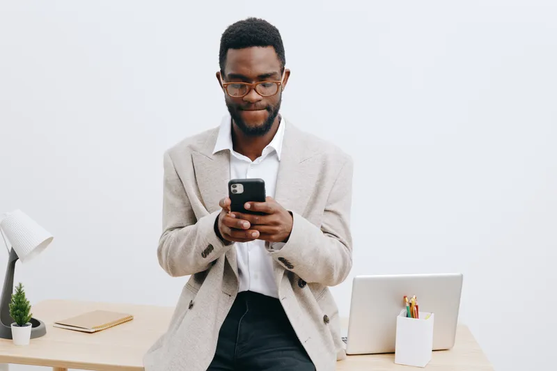 Confident young man of African descent using smartphone in modern workspace, wearing casual beige cardigan and glasses, standing by desk with laptop, white background, lifestyle and technology concept