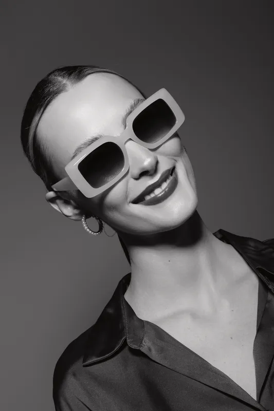 Smiling young Caucasian woman with fair skin wearing large sunglasses and hoop earrings, dressed in silk blouse, posing confidently in black and white studio portrait.