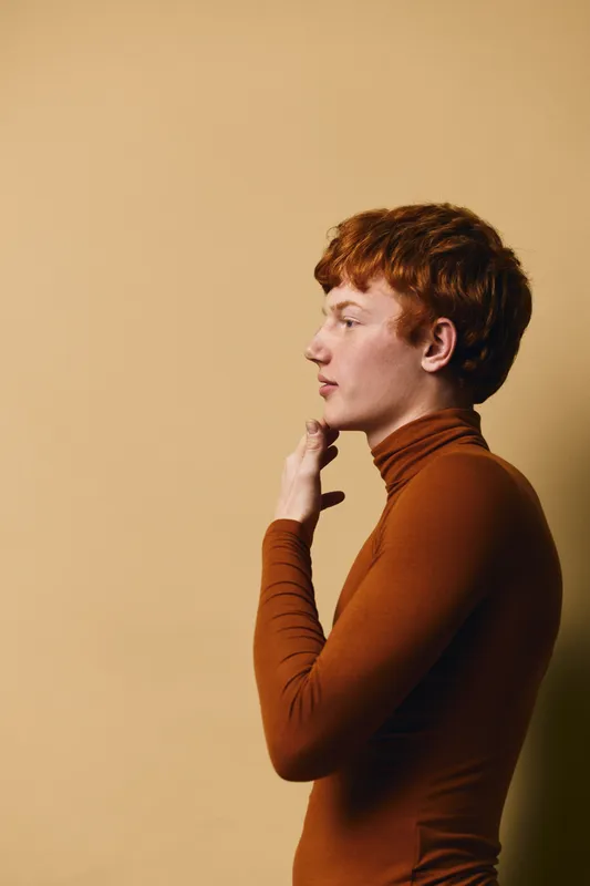 Profile of young man with red hair wearing brown turtleneck sweater standing thoughtful against beige background in studio for portrait and fashion concept.