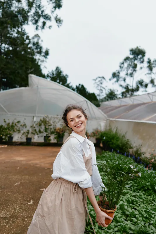 Young woman in a garden wearing an apron, holding a plant, smiling joyfully in a lush green backdrop, perfect for concepts like gardening and healthy living