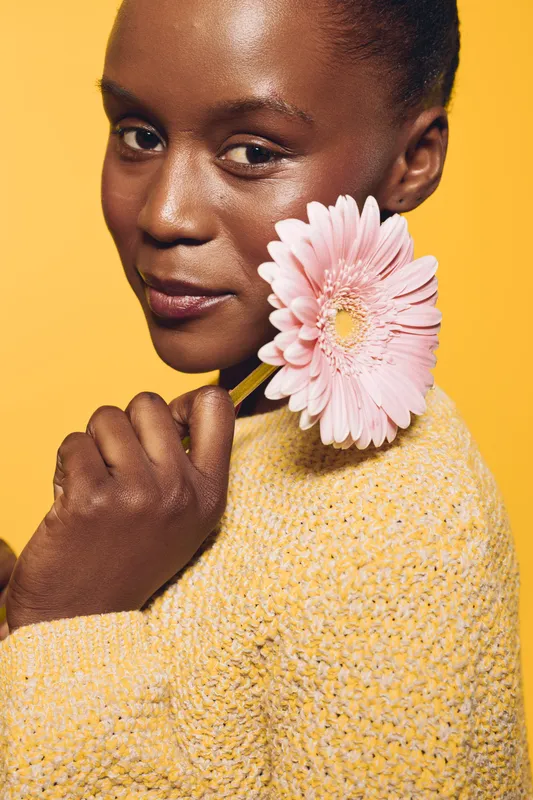Close-up of a smiling African woman with dark skin holding a pink gerbera flower near her face. She wears a yellow sweater against a bright yellow background. Concept for beauty and nature.