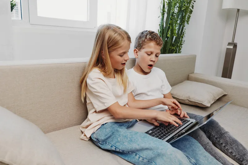 Two children sitting on a sofa using a laptop together in a bright living room. They appear focused and engaged, sharing a moment of learning and technology at home.