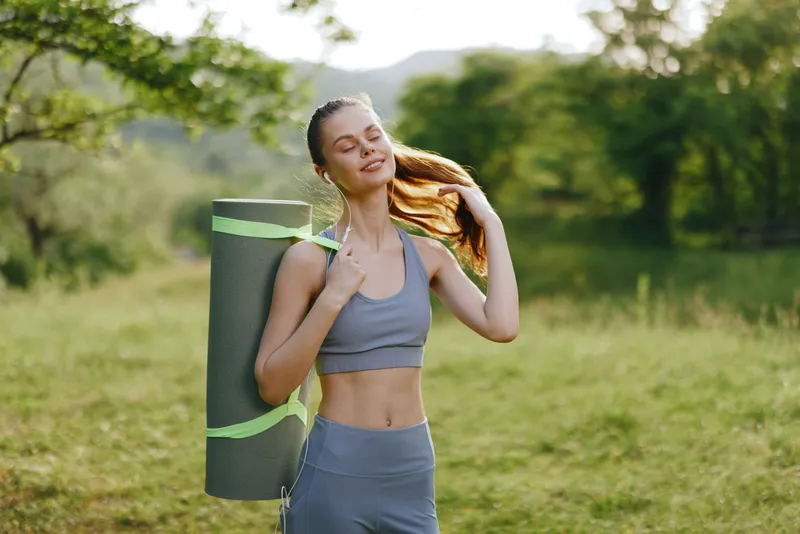 Young woman with a yoga mat enjoying nature, wearing sports attire, expressing joy in a green outdoor setting, embodying health and fitness