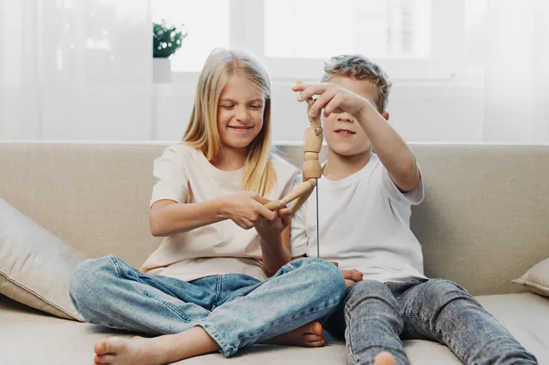 Two children sitting on sofa playing with wooden toy, smiling and having fun in bright cozy living room interior.