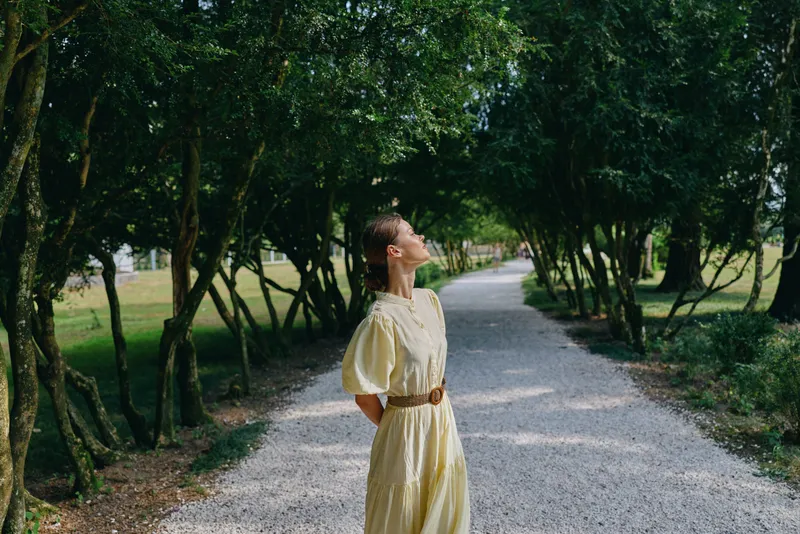 Woman in a yellow dress stands on a gravel path in a green park, looking up with a serene gaze, sunlight filtering through trees as nature creates a calm outdoor portrait