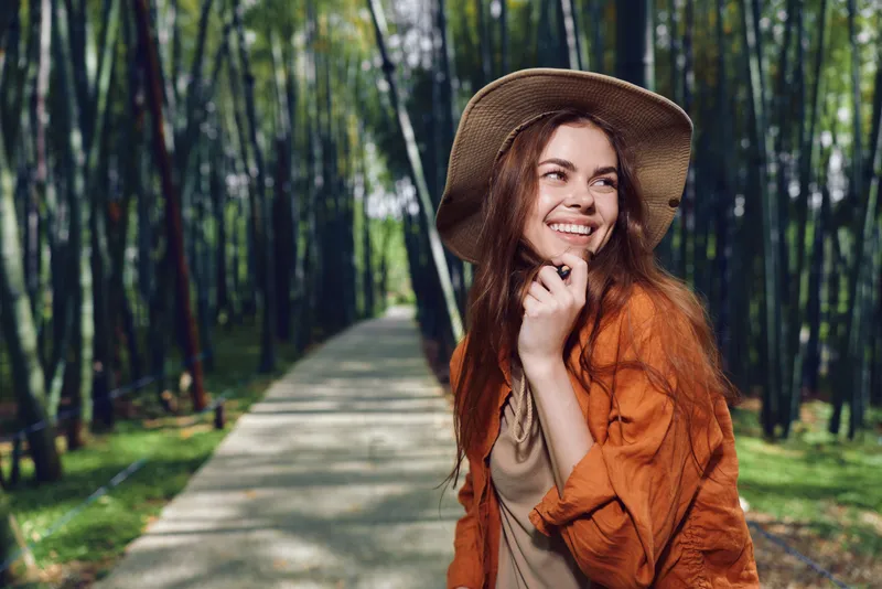 Woman smiling in a hat on a forest path, redhair and casual orange jacket, outdoor portrait of a happy traveler enjoying sunlight among tall bamboo trees and peaceful walkway.