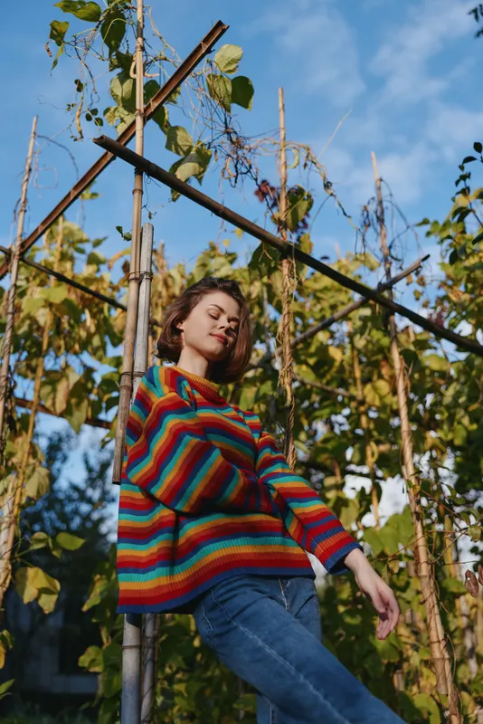 Woman in a rainbow sweater and jeans leaning on a wooden trellis in a garden, young adult smiling with closed eyes, relaxed pose, casual outfit, lifestyle fashion and sunny outdoor vibe.