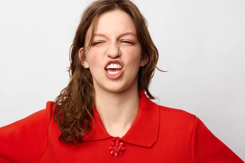 Young woman in red sweater makes funny face with eyes closed and mouth twisted. Studio portrait on white background. Concept of humor, expression, and casual fashion.