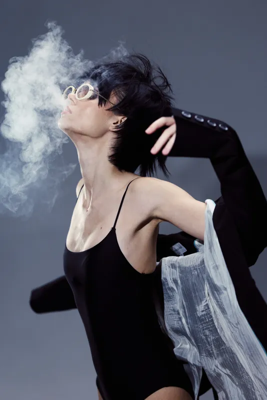 Stylish woman in black bodysuit exhaling smoke with dramatic lighting and modern fashion vibe.