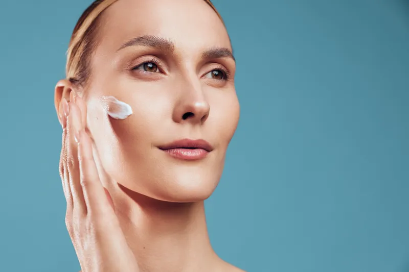 Skincare closeup of woman with face cream on cheek, calm expression, smooth skin, soft natural makeup, blue background, clean beauty mood, modern routine, hydrated