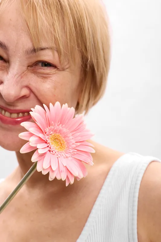 Woman portrait, flower pink daisy smile closeup elderly, blonde hair, soft studio light, face close to camera, white blouse, delicate petals, calm expression
