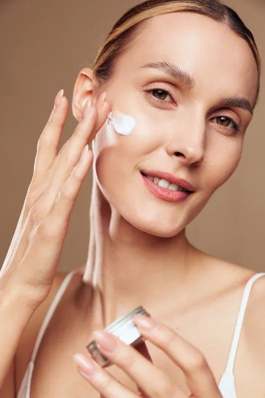 Woman applying face cream in closeup portrait, skincare care and wellness beauty, soft studio light on warm beige background, smooth hydrated skin