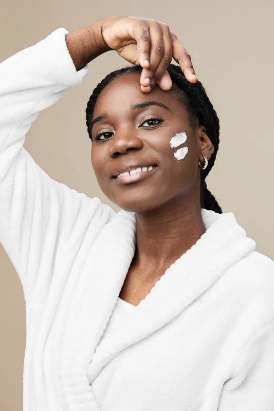 Young woman applying skincare product with a smile, showcasing healthy skin and confidence, dressed in cozy white robe against beige background