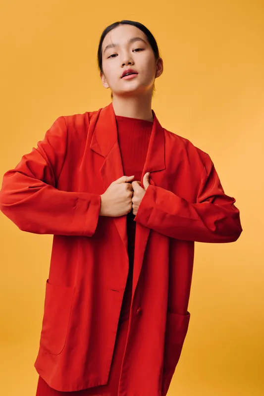 Young woman in red oversized jacket and matching outfit standing confidently against orange background with hands adjusting collar for fashion portrait and style concept.