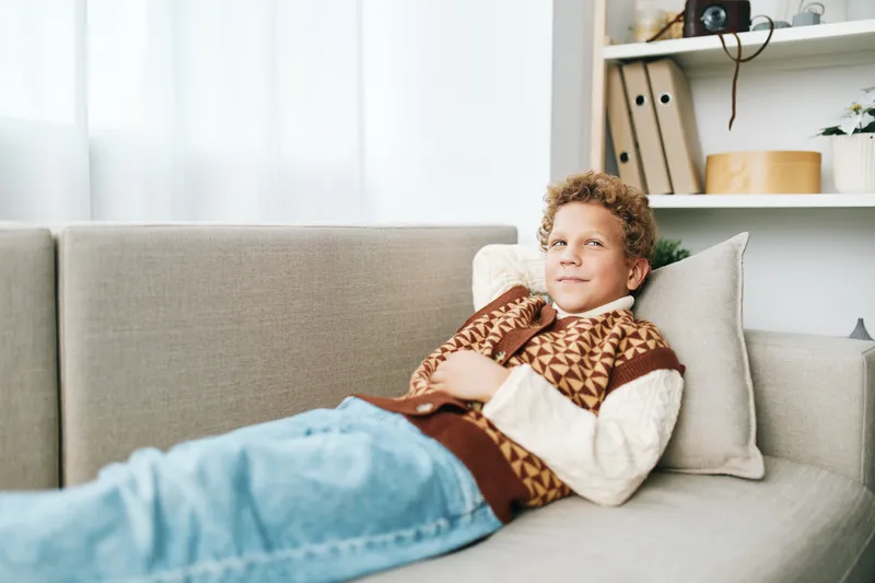 A young boy lounges on a light sofa wearing a patterned sweater and jeans, hands tucked under cheeks, gazing softly upward in a cozy, sunlit living room.