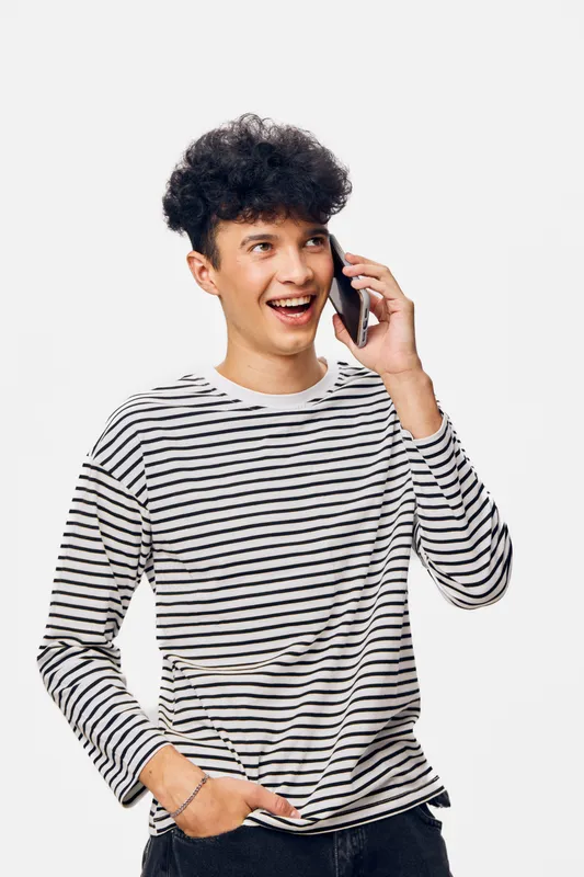smiling man talking on phone wearing striped long sleeve shirt standing on white background expressing happiness and engagement in conversation
