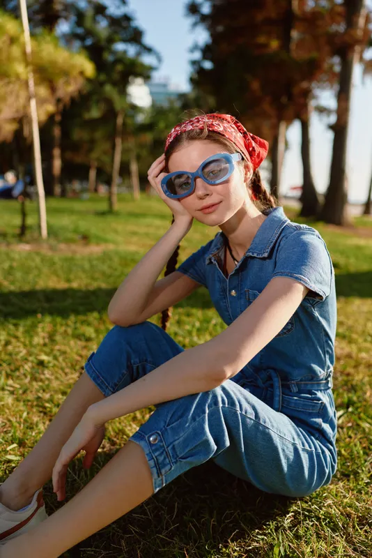 lifestyle woman in modern boho-western denim street style sitting outdoors on grass wearing blue sunglasses and red bandana, film color vibe, warm natural light