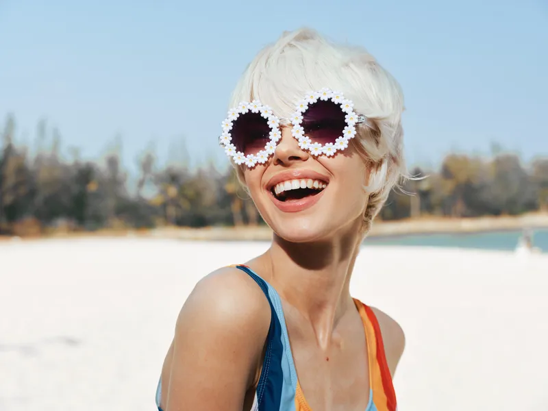 A bright portrait of a stylish woman at a sunny beach, wearing circular flower-framed sunglasses, radiant smile, and vibrant swimwear, capturing playful summer charm.