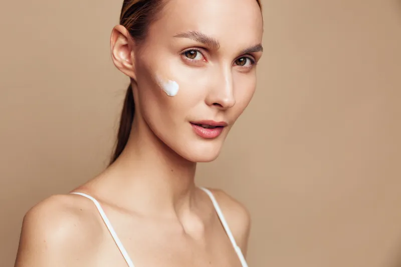 Young woman with clean skin applies moisturizing cream on her face looking at camera isolated on beige background in skincare beauty concept with natural makeup and fresh expression.