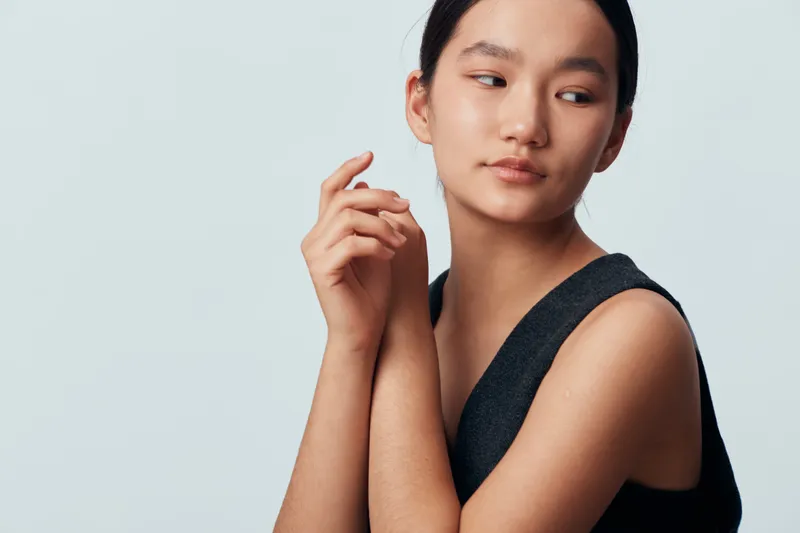 Young woman with calm expression posing with folded hands wearing sleeveless black top on plain light background, beauty portrait with natural makeup and soft lighting.