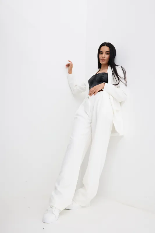 woman dressed in white suit leans against white wall, wearing black top and white sneakers, showing relaxed and stylish pose in minimalistic bright setting, modern fashion concept