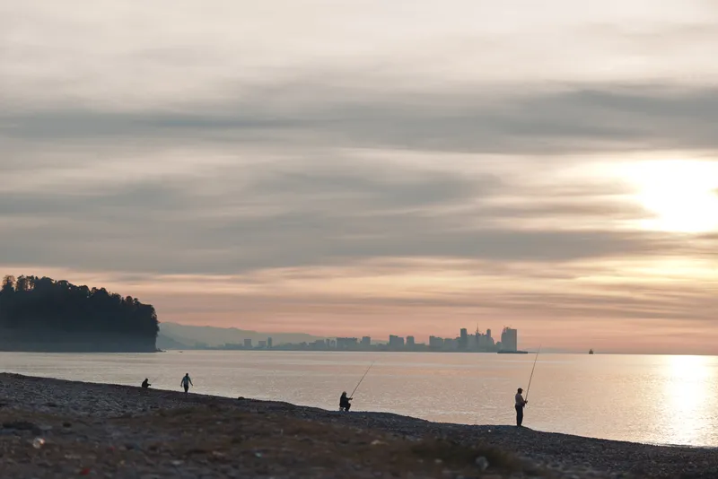 Beach at sunset with calm water and silhouettes of people fishing along the shore, distant city skyline on the horizon, warm sky reflecting on waves during golden hour.