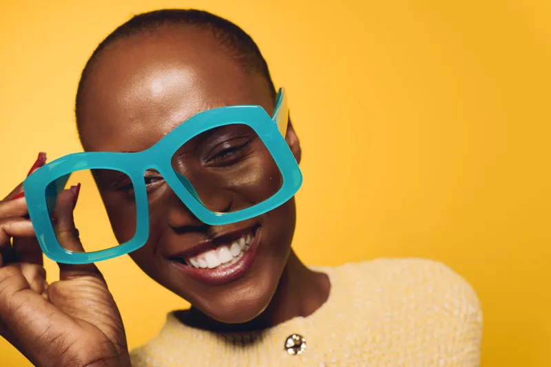 Smiling black woman with dark skin wearing oversized blue glasses and beige sweater on yellow background. Portrait of happy African female model with bright cheerful expression and joyful mood.