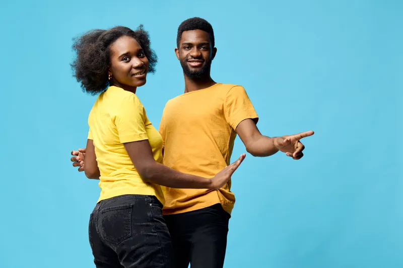 Young African couple dancing joyfully against a bright blue background, wearing matching yellow shirts, expressing happiness and unity through movement