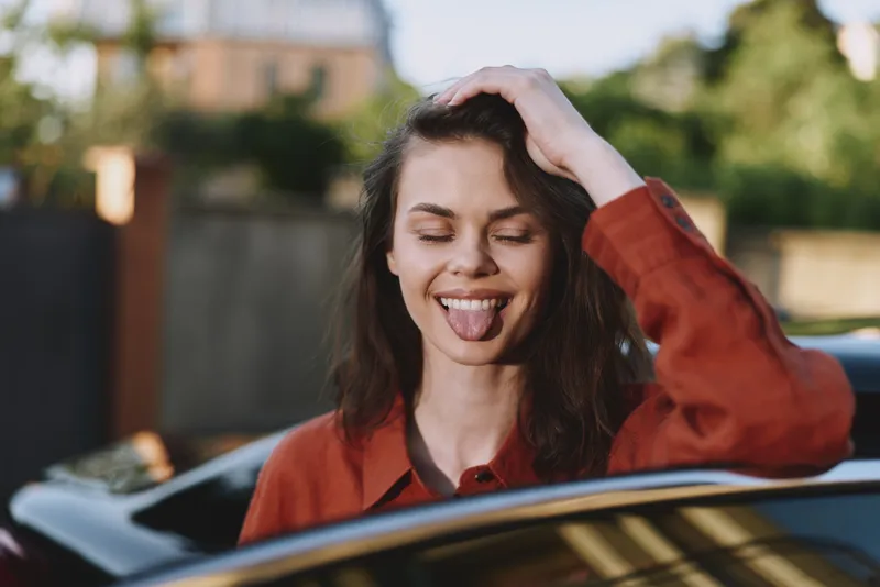 Playful woman with wavy hair wearing a red shirt leans out of a car window, sticking her tongue out. Bright outdoor light conveys carefree lifestyle and spontaneous mood