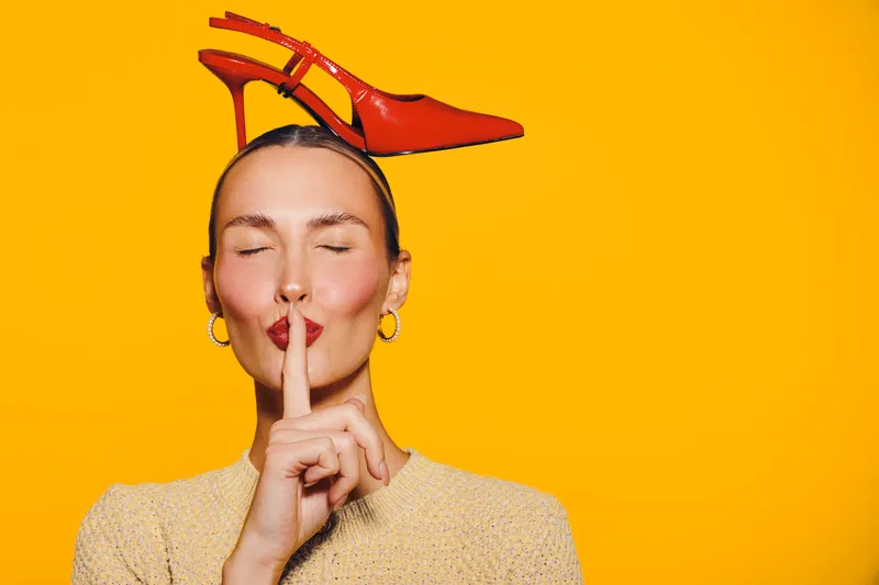 Young woman with closed eyes holds finger to lips in silence gesture. Red high heel shoe balanced on her head against bright yellow background. Concept of quiet, fashion, and creativity.