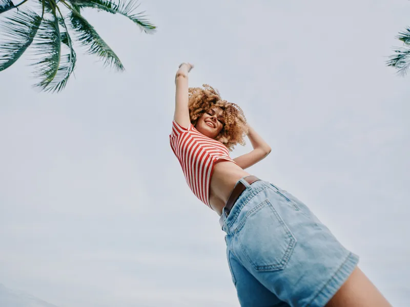 Playful portrait of a smiling young woman jumping outdoors, wearing a striped crop top and denim shorts, captured from a low angle against a bright sky.