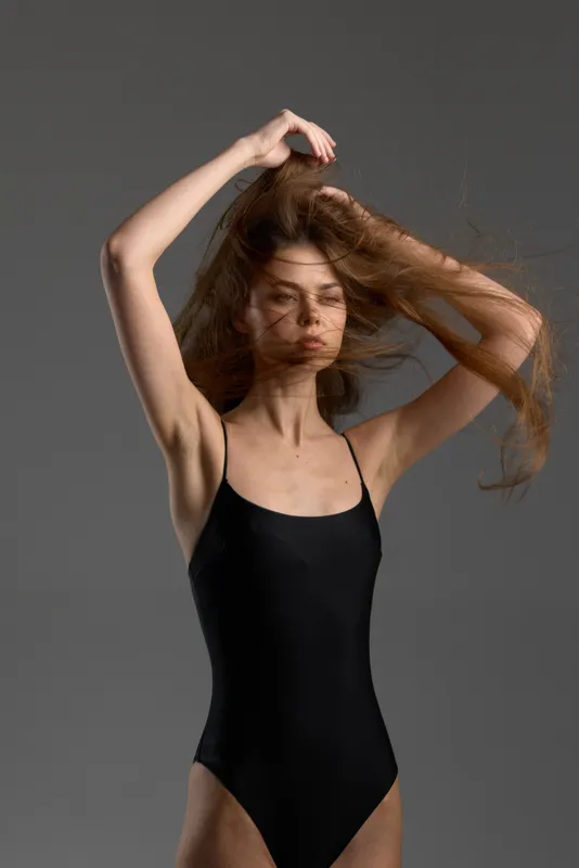 Young woman in black bodysuit poses with flowing hair against gray background, expressing motion and grace.