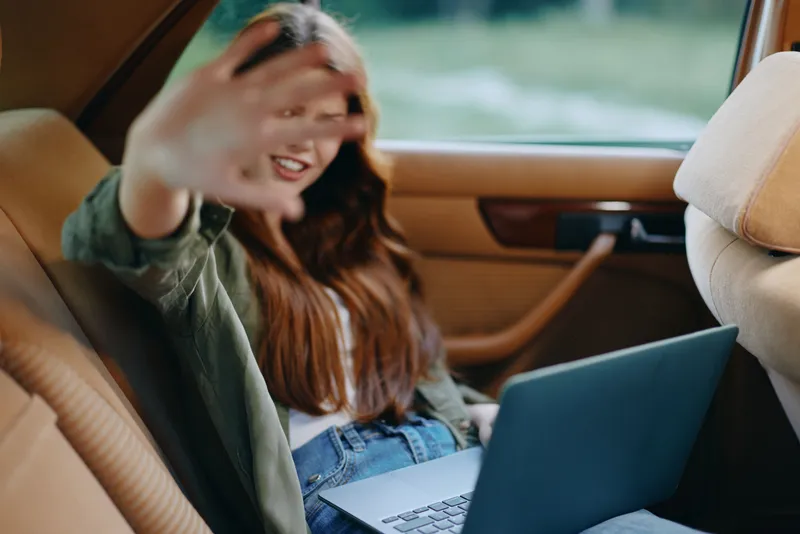 Young woman using a laptop in a car, smiling and playfully covering her face with her hand, showcasing a modern lifestyle and working remotely.